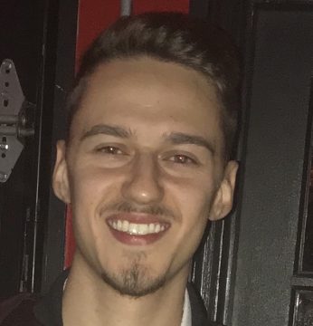 A young man with dark hair and a goatee smiles at the camera against a dark background.